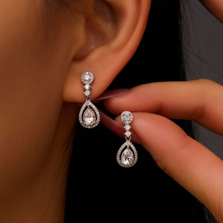 Teardrop-shaped diamond earrings being worn and held by a hand against a dark background